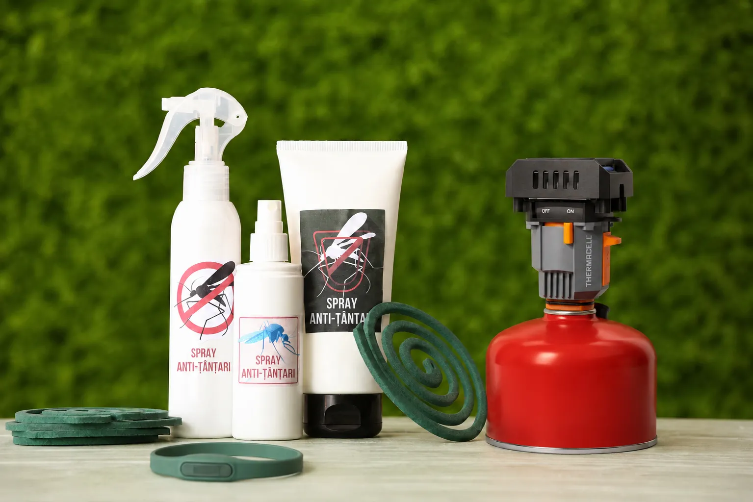 Spray-uri, spirale, brățară anti-țânțari și un dispozitiv Thermacell așezate pe o masă, în fața unui gard viu